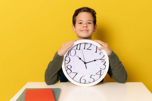 istock Student boy sitting on the desk holding big clock isolated over yellow background. 1365211868