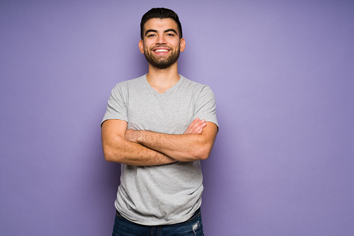 Handsome hispanic young man with casual clothes feeling confident and smiling in front of a purple studio background