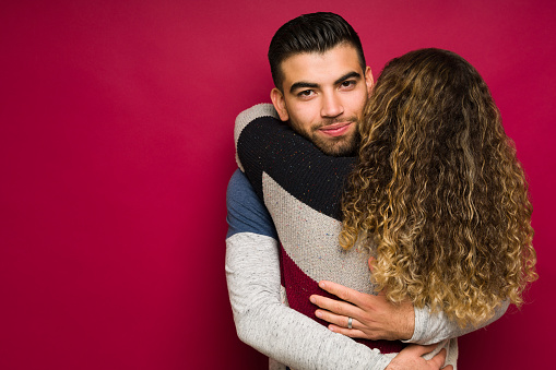 Portrait of a smiling young man hugging tight his beautiful girlfriend against a red studio background