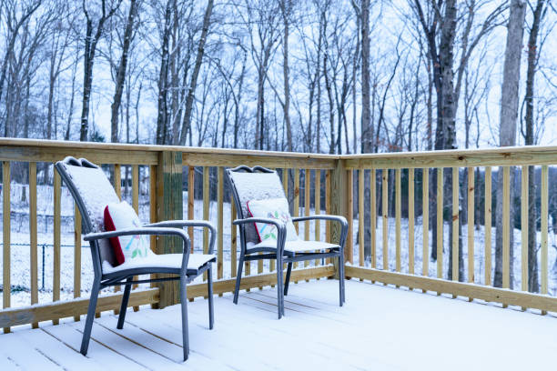 istock Two chairs, with pillows on them, sit on a wooden deck with fresh snow on them.  The background are forest trees and an overcast sky. 1365230382