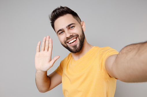 De cerca joven caucásico sonriente hombre guapo con camiseta básica amarilla casual haciendo selfie tomada en el teléfono móvil agitando la palma de la mano saludando a alguien aislado en un retrato de estudio de fondo gris. photo