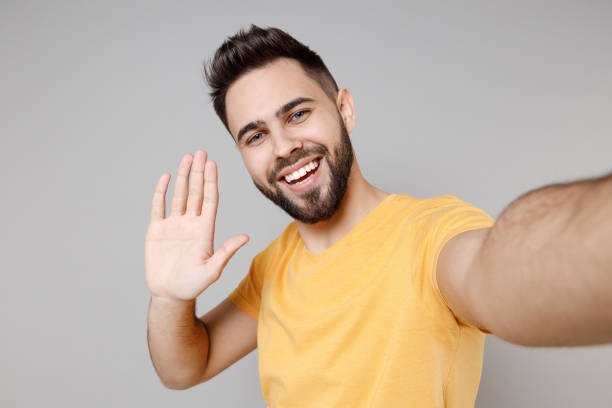 Close up young caucasian smiling bearded attractive man in casual yellow basic t-shirt doing selfie shot on mobile phone waving hand palm greeting someone isolated on grey background studio portrait.