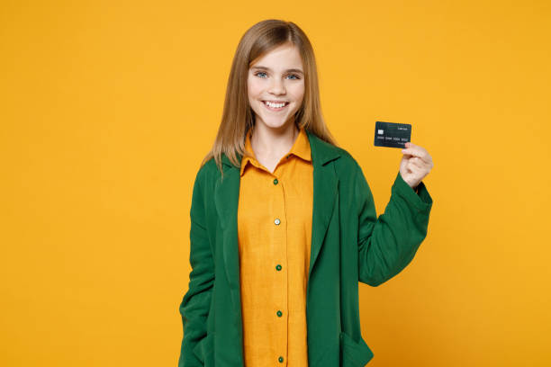 little blonde pretty nice kid girl 12-13 years old wear casual clothes green shirt hold credit bank card isolated on yellow orange background children studio portrait. childhood lifestyle concept - teenager 14 15 years 13 14 years cheerful imagens e fotografias de stock