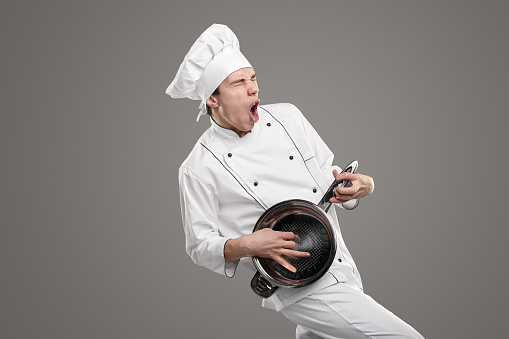Funny young male cook in white uniform and hat pretending to play musical instrument and sing while holding metal frying pan against gray background