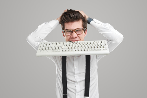 Overworked male coder in glasses biting keyboard and tearing hair on gray background in studio