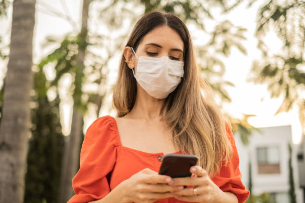 istock Mid adult latin woman using the mobile phone outdoors - using a face mask 1365256593