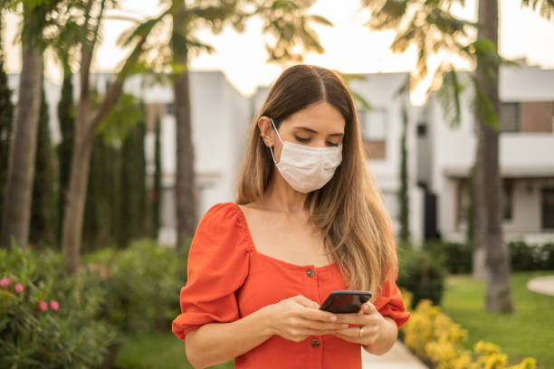 istock Mid adult latin woman using the mobile phone outdoors - using a face mask 1365256612
