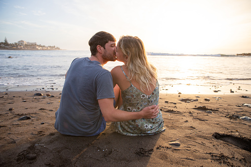 Couple kissing while sitting on the beach and watch the sunset, lovers are hugging. Traveling couple on beach during their vacation