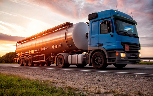 A blue modern truck with a semi-trailer tanker transports dangerous goods against the backdrop of the evening sunset. The concept of transportation of liquid cargo, hazard class ADR.