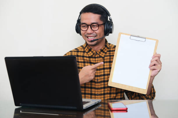 istock Adult Asian man wearing headset and showing empty white paper when doing online meeting using laptop 1365293743
