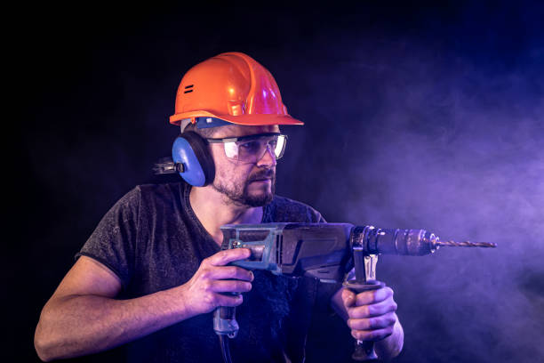 A young man in overalls protective helmet hard hat hold electric drill isolated on dark background in studio. Instruments accessories for renovation apartment room. Repair home concept.