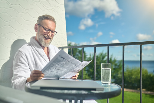 Smiling happy man in the white bathrobe and spectacles seated at the glass coffee table on the balcony