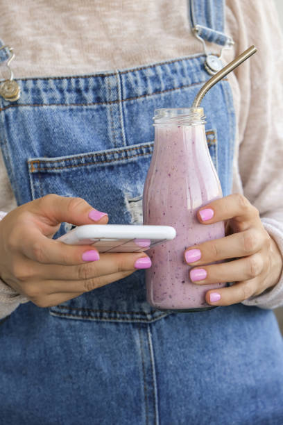 female hands holding blueberry smoothie topped with blueberries. woman drinking glass of breakfast protein smoothie drink. using mobile phone, social media, snack break, online studying, recipe - blueberry smoothie milk shake drink imagens e fotografias de stock