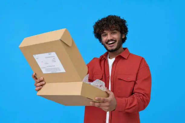 Photo of Happy indian young man holding open parcel box isolated on blue background.