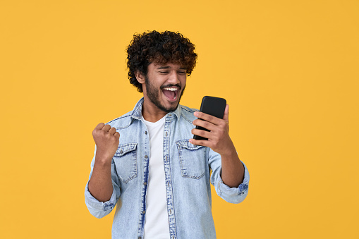 Excited young indian man winner using smartphone isolated on yellow background.