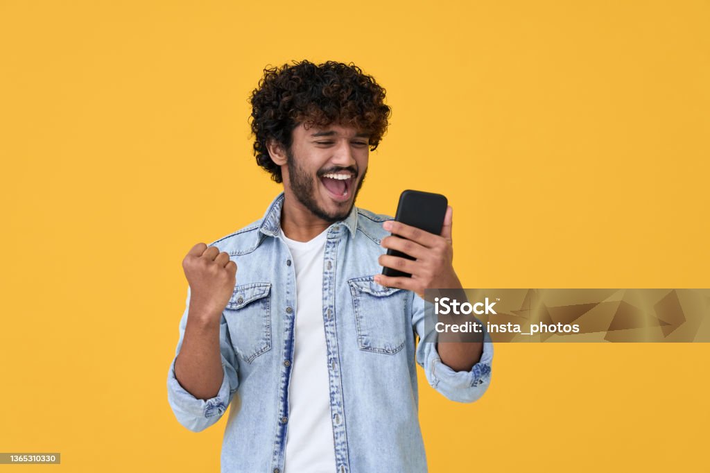 Excited young indian man winner using smartphone isolated on yellow background. Excited happy young indian man winner feeling joy using smartphone winning lottery game, betting, getting cashback online gift in mobile app message holding cell phone isolated on yellow background. Happiness Stock Photo