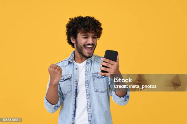 Excited Young Indian Man Winner Using Smartphone Isolated On Yellow Background Stock Photo - Download Image Now