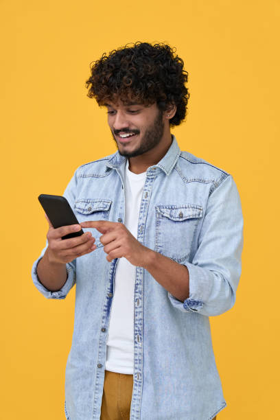 Happy indian young man using cell phone isolated on yellow background. Happy indian young man using cell phone isolated on yellow background. Smiling ethnic hipster guy holding smartphone playing game in app, dating buying online in ecommerce store on cellphone. Vertical person paying stock pictures, royalty-free photos & images