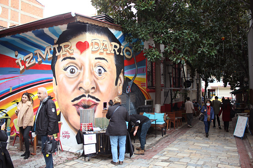 Izmir, Turkey - December 11, 2021 : Dario Moreno street view with Moreno portrait painting on building's wall. People walking on the touristic street, some posing for photo,  sitting at the cafe. Moreno was famous singer from Izmir in Turkey.