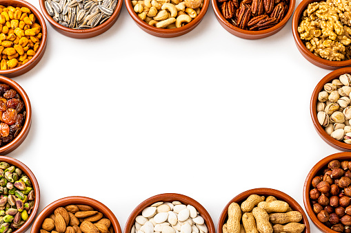 Top view of several bowls disposed at the borders of the image leaving a useful copy space at the center on a white background. Each bowl is filled with a different nut. The composition includes pistachios, walnuts, hazelnuts, cashews, almonds, sunflower seeds, pumpkin seeds, pecans, raisins and peanuts.