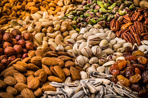 High angle view of a background made of various scattered nuts. The composition includes pistachios, walnuts, hazelnuts, cashews, almonds, sunflower seeds, pumpkin seeds, pecans, raisins and peanuts.