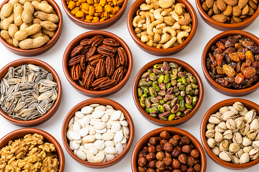 Top view of a background made of several bowls each filled with a different nut. The composition includes pistachios, walnuts, hazelnuts, cashews, almonds, sunflower seeds, pumpkin seeds, pecans, raisins and peanuts. The bowls are on white background