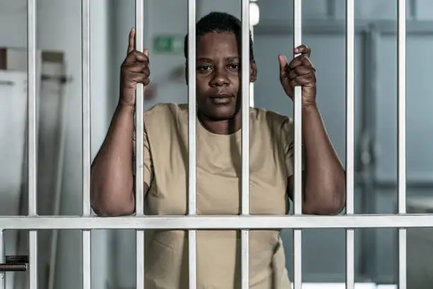 Photo of Young afro woman looking  serious and desperate   behind bars which may be prison bars or those of a security gate