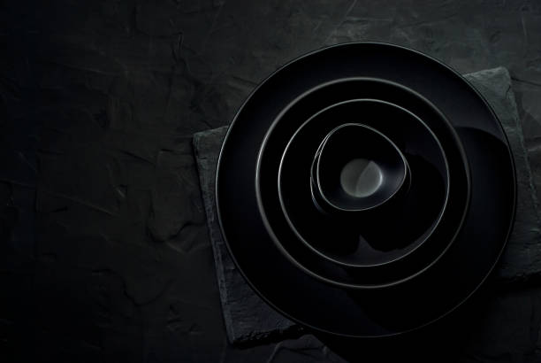 istock Black tableware on a dark background. Plates and bowls of different shapes and sizes are stacked in a pile. Contrasting dramatic light as an artistic effect. 1365331449