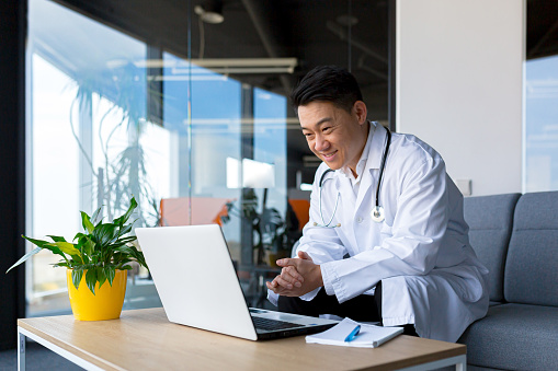 Médico asiático amigable usa computadora portátil para comunicarse de forma remota con el paciente, el hombre en la clínica consulta en línea, mira la pantalla del monitor y la cámara web photo