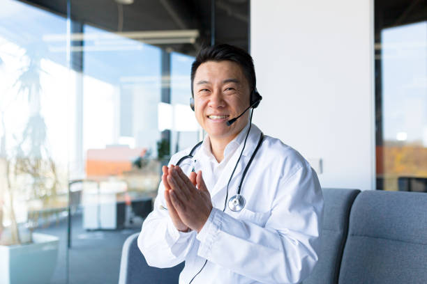 porträt eines erfahrenen asiatischen arztes, der in die kamera schaut und spaß daran hat, mit dem patienten zu sprechen, freundlich zu lächeln und sich über die genesung des patienten zu freuen, aus der ferne zu arbeiten, videoanruf mit headset - amicably stock-fotos und bilder