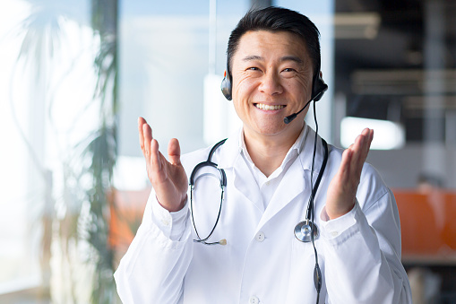Portrait of an experienced Asian doctor, looking at the camera and having fun talking to the patient, smiling amicably and rejoicing in the patient's recovery, working remotely, video call with headset