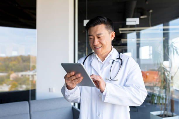 asiático alegre e amigável em uma clínica moderna, chamada de vídeo online, usa um tablet, para se comunicar com os pacientes, e reuniões online com colegas - doctor digital display digital tablet healthcare and medicine - fotografias e filmes do acervo