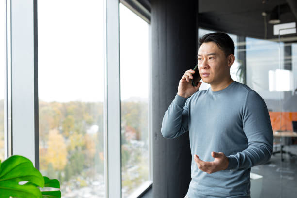 hombre serio y enojado hablando por teléfono en la oficina, asiático pensativo en la ventana enojado - quejándose fotografías e imágenes de stock