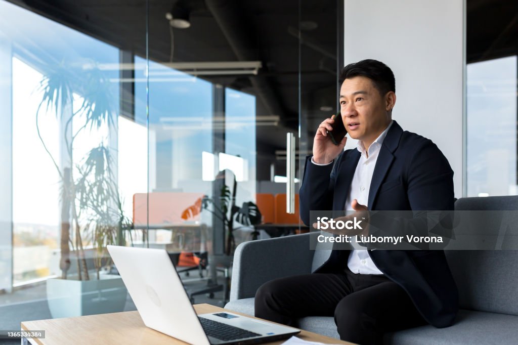 Thinking asian businessman businessman, talking on the phone working in a modern office, listening seriously Using Phone Stock Photo