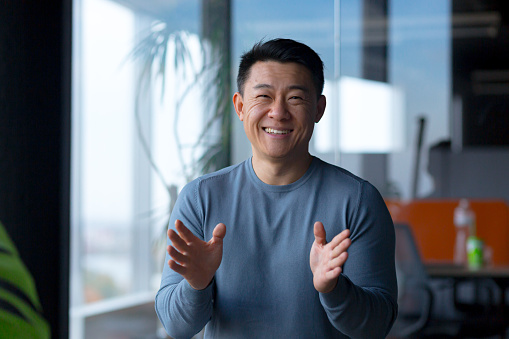 Portrait of a cheerful employee, looking at the camera and gesturing with his hands, Asian communicates on a video call, looks at the webcam