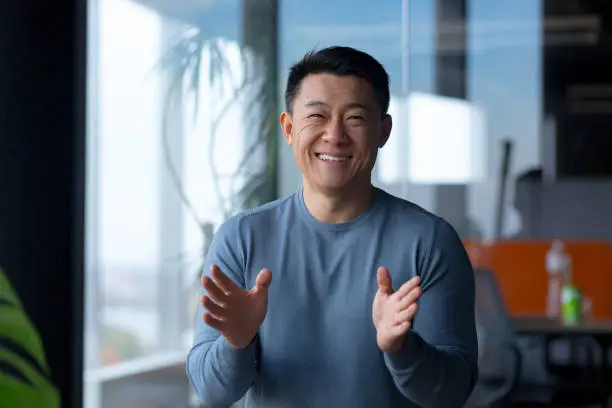 Photo of Portrait of a cheerful employee, looking at the camera and gesturing with his hands, Asian communicates on a video call, looks at the webcam