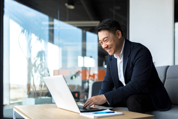 hombre de negocios asiático senior que trabaja en una computadora portátil en línea, sonriente y regocijado, feliz jefe propietario de negocios que trabaja en la oficina - japonés oriental fotos fotografías e imágenes de stock