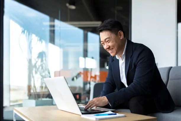 Photo of Senior Asian business man working on laptop online, smiling and rejoicing, happy boss business owner working in office