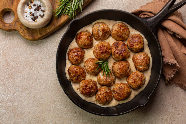 Homemade Swedish Meatballs made with ground meat, onion, egg, bread crumbs and nutmeg. With creamy gravy in black pan skillet.  On beige concrete table. stock photo