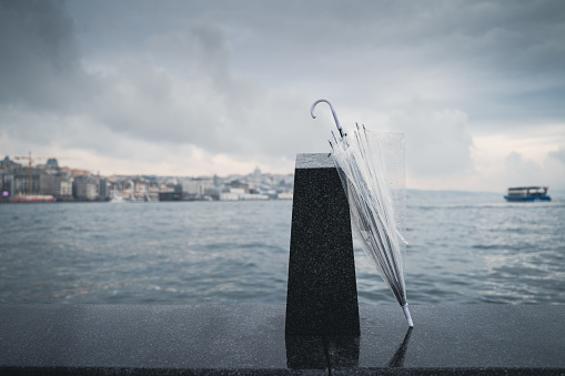 front view of closed transparent umbrella leaning against a black stone block near water with clouds above and shoreline in the background