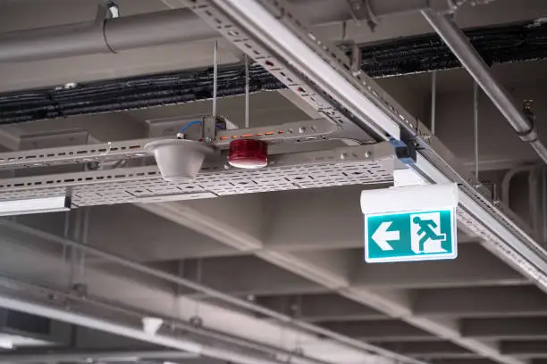 front view closeup of lighted green exit sign hanging on building pipes and pointing the way out of an industrial building