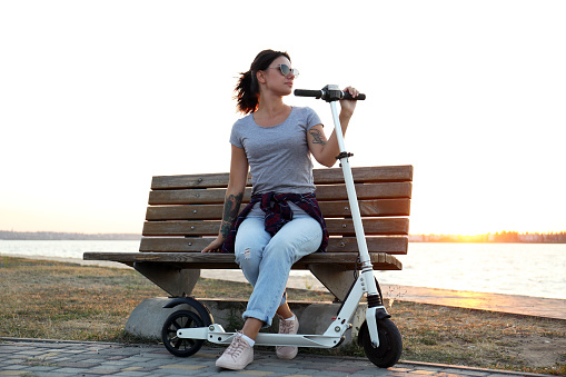 Young woman with kick scooter sitting on bench outdoors