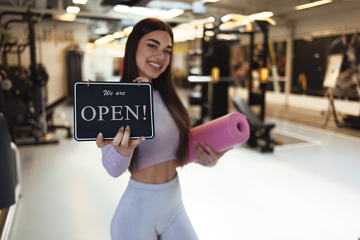 A beautiful young girl, looking at the camera with a smile on her face, holds a sign that the gym has reopened after a long time. A young handsome woman holding a yoga mat indicates that the gym has reopened.