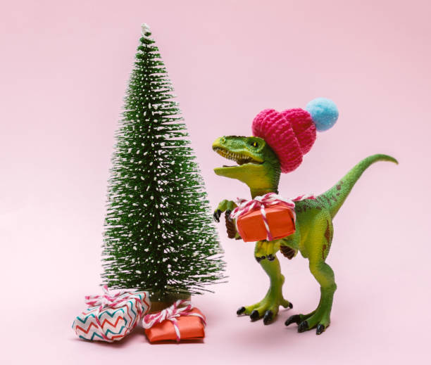 istock Funny plastic toy dinosaur wearing knitted hat with  present boxes and evergreen tree on a pink background. 1365358338