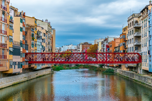 Red metal Eiffel bridge over the Onyar river in Girona, Spain. Cityscape with beautiful old buildings on the embankment and their reflections in the water