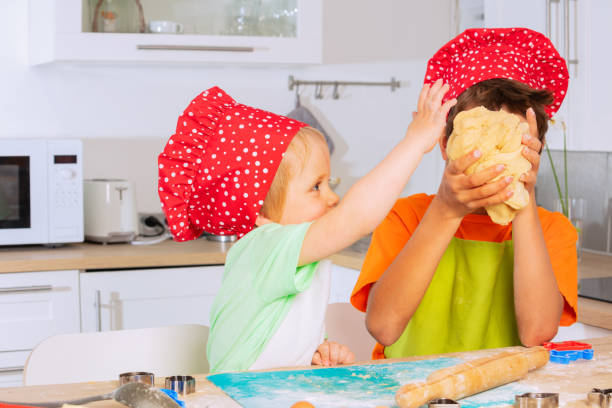 Toddler boy play with brother who crumble dough using hands wearing chef red hat and smiling