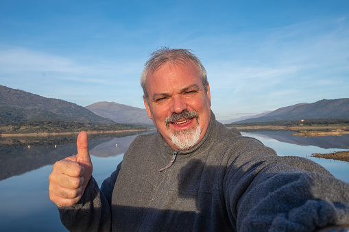 Mature hiker taking a selfie portrait on top of the mountain next to a lake, happy senior smiling at camera, healthy lifestyle, Hiking exercise and health
