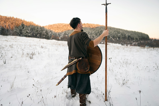 Rear view of unrecognizable mid-adult Caucasian man, wearing the warrior costume, shield and spear, at nature during winter sunset