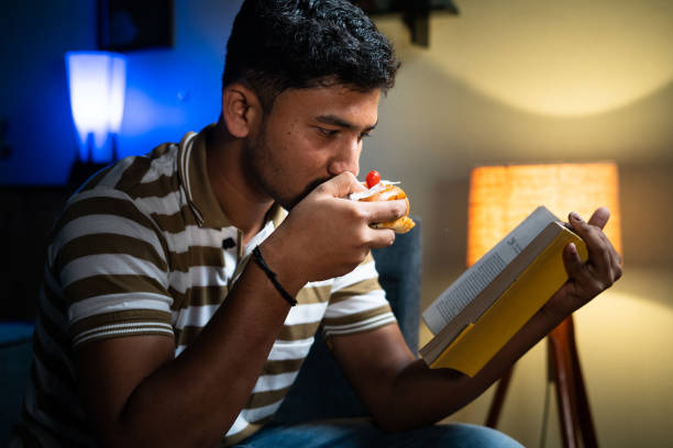 young indian student eating unhealthy food while reading book at home - concept of unhealthy lifestyle, hobby, skill and knowladge development - eating sandwich emotional stress food imagens e fotografias de stock