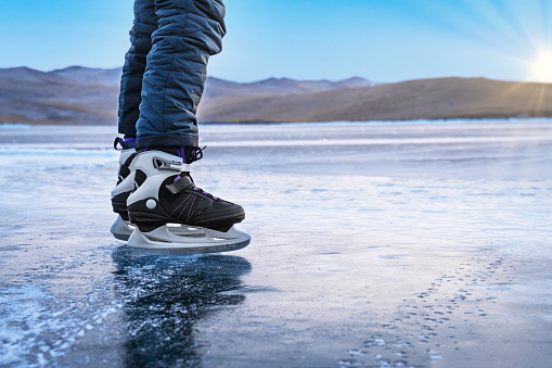 Fun ice skating on a frozen lake on a sunny winter day. View of the beautiful transparent blue ice of frozen Baikal Lake. Human in skates travels on the ice. Winter outdoor activities.